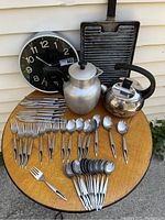 Full view of mid-century kitchen items on round wood table including cutlery set, coffee pot, kettle, wall clock, and grill pan.