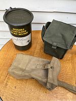 Three military items displayed on wooden surface: metal canister, green canvas bag, and tan canvas satchel with stamps inside flap