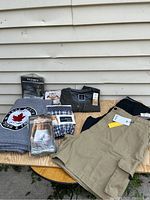 Full lot viewed from the front on wood table outside against siding wall, khaki shorts, green shirt, black shirt, fleece blanket, packaged briefs visible.