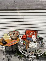 Photo of the full lot showing the various vintage kitchenware items arranged on two tables outdoors against a white siding wall. Includes teapot, floral plates, tins, plastic boxes, utensils.