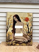Full view of needlepoint tapestry showing nude kneeling woman and nature background, framed and leaning against siding.