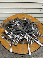 Pile of assorted stainless steel forks, knives, and some spoons on a round wooden table, outdoor setup.