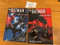 Two Batman Under The Hood graphic novels volume 1 and 2 on wooden surface, front covers visible, volume 1 in blue and volume 2 in red.