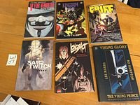 Front covers of six graphic novels including V for Vendetta, Kingdom Come, Grief, San and Twitch, Lovelost, and Viking Glory placed on a wooden surface.