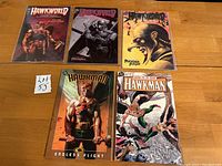 Front cover view of five graphic novels arranged on a wooden surface, showing artwork and title logos