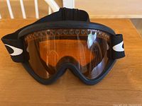 Front view of Oakley ski goggles with orange-tinted lenses and black frame on wooden table.