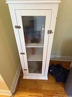 Front view of small white wooden cabinet with ribbed glass door showing two interior shelves and metal latch.