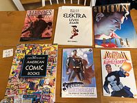Front covers of six comic and graphic novels on a wooden table, showing titles and artwork.