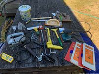 Overview of various tools spread on wooden table including electric jigsaw, saw, scrapers, sanding blocks, pliers, glue gun, and tape.
