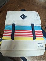 Front view of the Herschel backpack showing cream canvas material, multicolor horizontal stripes, black magnetic straps, and Herschel logo patch.