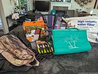 Photo showing all items arranged on furniture: wireless meat thermometer set in box, picnic blanket green with palm tree print, camo toiletry bag, blue and white floral insulated lunch bag, high volume water filter kit in large packaging, and six black clips.