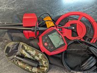 Red and black metal detector with coil, headphones, and handheld probe laid out to show included accessories.