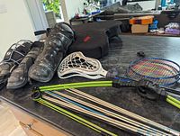 Photo showing a variety of sports equipment including black riding boots, lacrosse stick, protective pads, badminton rackets, and longbow with wooden arrows