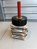 Stack of 123 assorted 45 RPM records with paper sleeves and loose discs on a red spindle.