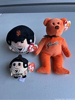Photo showing three SF Giants Beanie Babies: an orange bear with Giants embroidered on chest and two baseball player dolls in helmets and uniforms, all with attached TY heart tags.