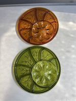 Photo showing two colored collectible glass serving dishes, one amber and one green, both with leaf and segmented designs.