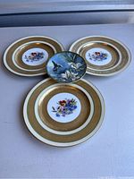 Four collectible plates displayed together: three gold flowered floral design plates and one smaller bird-themed plate with hummingbird and lilies.