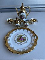 Full view of 24 karat gold accented Bavaria tea set including teapot, sugar bowl with lid, creamer, two teacups, and large decorative tray. Each piece features a central painted scene of figures in classical attire, surrounded by gold filigree.