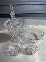 Four clear cut glass pieces on a surface, highlighted by natural light: a lidded candy dish, a fluted bowl, a small bowl, and a creamer.