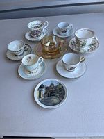 Collection of eight assorted teacups with matching saucers and one decorative plate arranged on beige table.