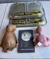 Two sealed Marsh Allen E.T. TV trays stacked; front tray with some scratches and torn wrapping; two E.T. plush dolls and a book placed beside trays on white table.