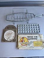 Metal fish-shaped griller basket, ceramic bread/bun warmer with lid, vintage 36-count ravioli mould with original box arranged on a flat surface.