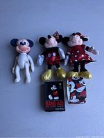 Photo shows two Beanie Babies plush toys (Mickey Mouse and Minnie Mouse) with tags attached, one vintage Mickey Mouse doll, unopened box of Mickey Mouse Band-Aids, and a roll of Mickey Mouse duct tape, all arranged on a blue surface.