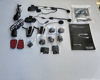 Full layout of the two Cardo Packtalk Duo headset units with wired microphones, speakers, multiple small bags of mounting hardware, clamps, red accessories, user guide, and cables.