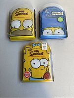 Photo shows three limited edition boxed DVD sets of The Simpsons featuring complete seasons 6, 7, and 8. Each box features a different Simpsons character: Homer for season 6, Marge for season 7, and Bart for season 8.