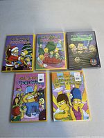 Five DVD cases of The Simpsons episodes lying on a flat surface. Four are sealed in plastic wrap; one is unsealed. Covers feature colorful artwork depicting characters in themed poses.