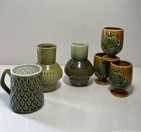 Front angle view of two ceramic vases, three brown pedestal cups with green apple motifs, and one green and brown geometric patterned ceramic mug.
