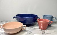 Full view of four vintage ceramic dinnerware pieces: a large blue bowl with handles, a peach-colored two-handled bowl, a red conical mug, and a blue mug. Arranged on marble surface against plain background.