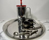 Stainless stovetop coffee press with red handle and knob, two dark glaze espresso cups, four stainless coffee spoons on large round stainless steel tray