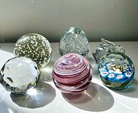 Six different glass paperweights arranged in a row showing their shapes, colors, and bubble designs under bright light.