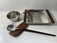 Photo showing the four serving pieces: stainless steel bowl with wooden handles, rectangular stainless steel butter warmer with wooden handles, wood and stainless steel spoon, and rectangular marble and wood cheeseboard.