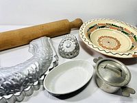 Wooden rolling pin, decorative ceramic plate, metal baking molds, and two ceramic dishes laid out on a white surface.