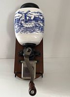 Front view of the wall-mounted coffee grinder showing ceramic container with blue dutch windmill design, metal handle, and wooden base.
