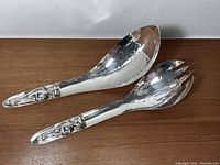 Pair of hammered silver-plated salad servers with detailed First Nations Kwakiutl totem face handles shown on wooden surface.