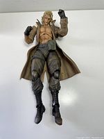 Full body front view of Liquid Snake figurine lying on flat surface, showing open tan coat, muscular torso, and black boots.