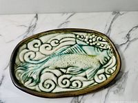 Top view of oval studio pottery dish showing fish and swirling wave relief glazed in green and brown tones on white base with crackle glaze.