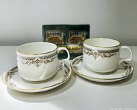 Two Royal Doulton Wardair cups and saucers with gold and brown Repton pattern along edges. Two Eaton's playing card decks visible behind them, one unopened in wrapping.