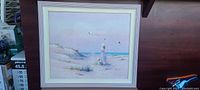Framed artwork showing a beach scene with two figures, one woman, one child, standing on sand dunes by the ocean, set in a white wooden frame, minor frame wear visible.