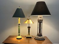 Three lamps on a wooden surface against a plain wall: a green ceramic lamp, a brass Orient Express lamp without chimney, and a large lamp with a classical column base and black shade.