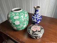 Three decorative pieces displayed on wooden surface: blue and white cloisonné vase, green ceramic vase with white blossoms, black floral trinket box.