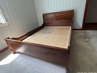 Slightly angled front-side view of the wooden sleigh bed frame showing curved headboard, footboard and side rails, on carpeted floor.
