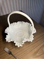 Large Fenton milk glass basket with ruffled edge and hobnail pattern from an angled top-down view on wooden table alongside a fork for size comparison.