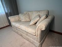 Front view of light-colored floral patterned love seat with three cushions, showing solid frame and table lamp beside it.