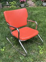 Front angled view of mid century child's rocking chair with orange vinyl seat and backrest on metal rocker frame placed outdoors on grass.