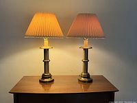Two brass table lamps on a wooden surface, lit, showing pleated fabric shades, one with repaired pleat