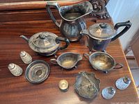 Photo of various silver plate serving pieces including a large hammered water pitcher, teapot, sugar bowl, small bowls, and salt and pepper shakers arranged on a wooden surface.